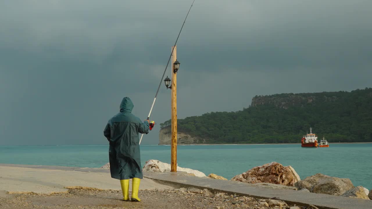 一个穿着蓝色雨衣和黄色靴子的渔夫站在暴风雨中的海上，手里拿着一根鱼竿。巨浪冲击着附近的岩石。在远处，一艘船和群山在海湾。