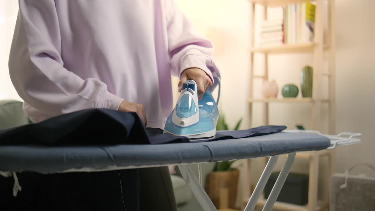 一名亚洲妇女熨衣服，准备外出办事。黄昏时分，阳光透过窗户照进来。