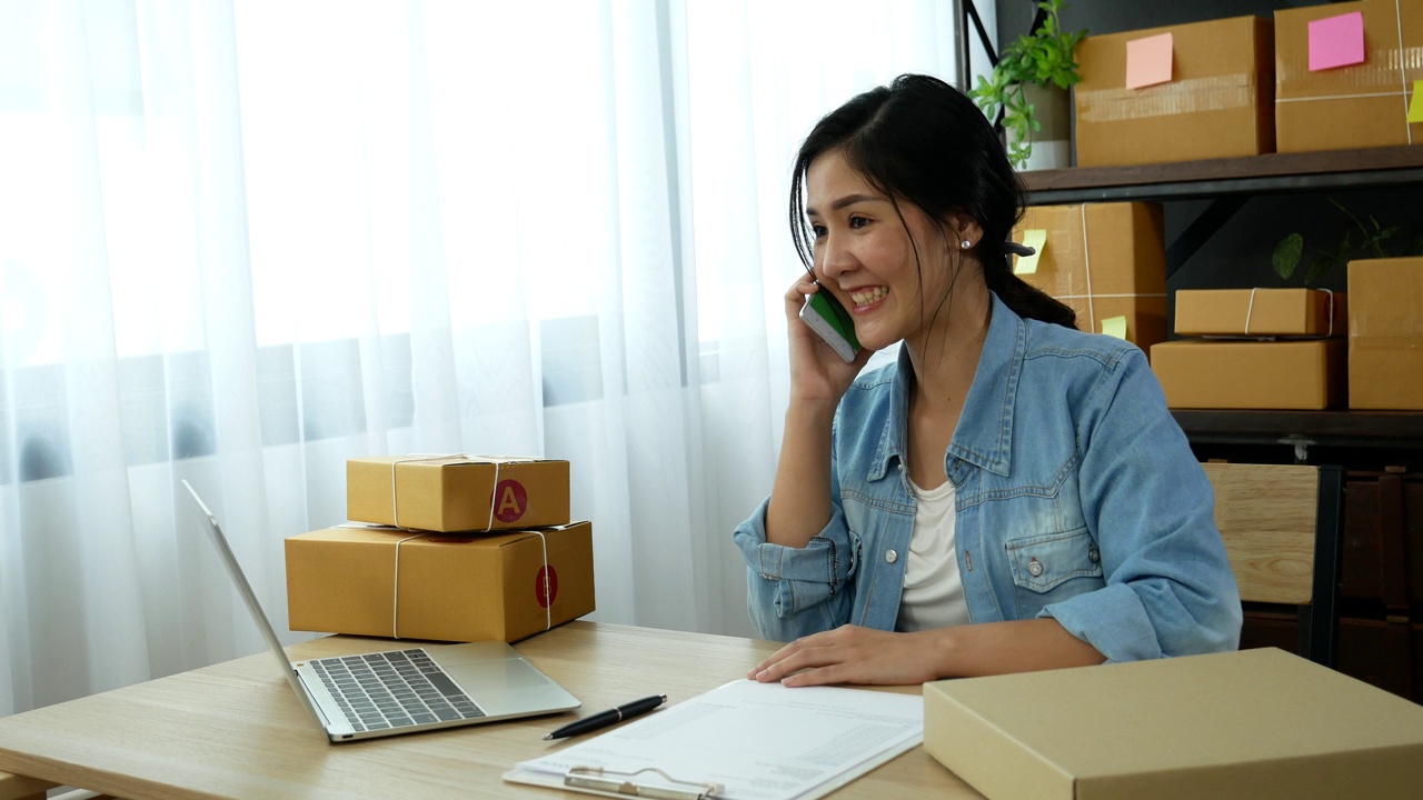 网上卖家近距离接触年轻的亚洲女子，她正在输入笔记本电脑键盘，检查网上订单，检查货物库存，发货包裹，发货邮政。亚洲女性在家办公室做小生意