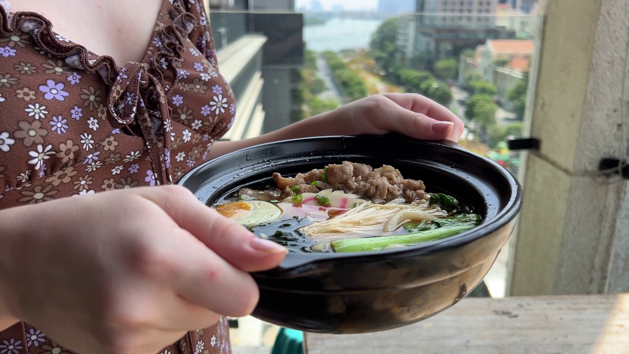 一个女孩手里拿着热汤，黑色的碗里盛着亚洲面汤、鸡肉、豆腐、蔬菜和鸡蛋的拉面。板岩的背景。前视图。