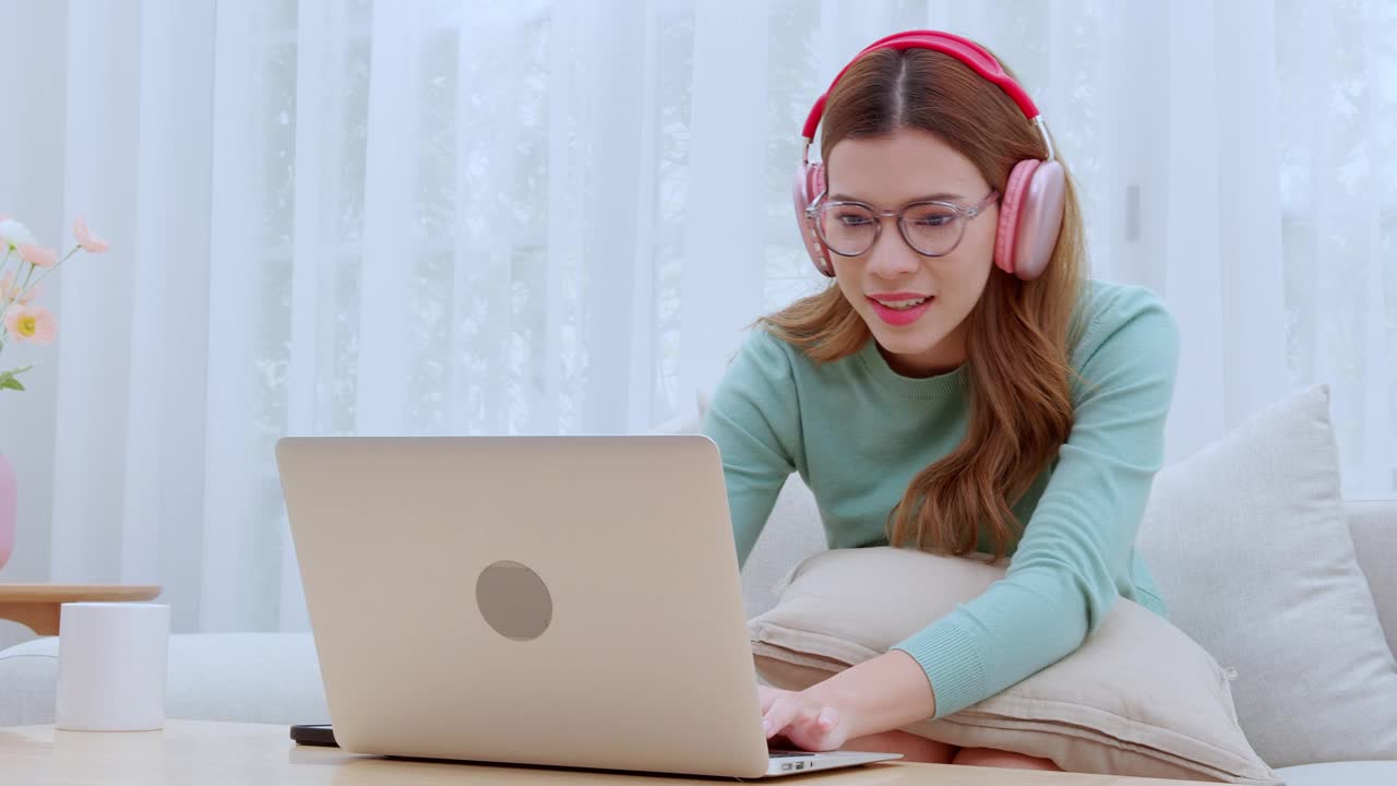 年轻的亚洲女性在家里的客厅里戴着耳机欣赏音乐，在笔记本电脑上工作，戴着耳机的女性使用笔记本电脑，远程办公或视频通话，数字游牧。