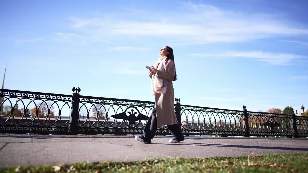 一名全身女子走在城市中心的水岸上，对着蓝天，用手机交流、通信或阅读信息。秋天在户外悠闲地漫步大自然。