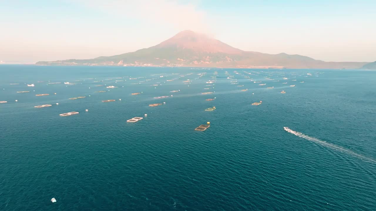 鸟瞰图，一艘渔船在鹿儿岛湾的蓝色海水和鱼笼之间驶向樱岛火山