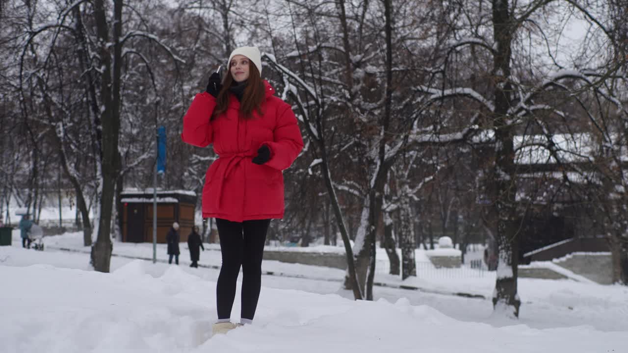 无忧无虑的年轻女子全身画像，穿着温暖的冬衣，在城市公园玩雪，用智能手机聊天。快乐的女性通话细胞享受谈话