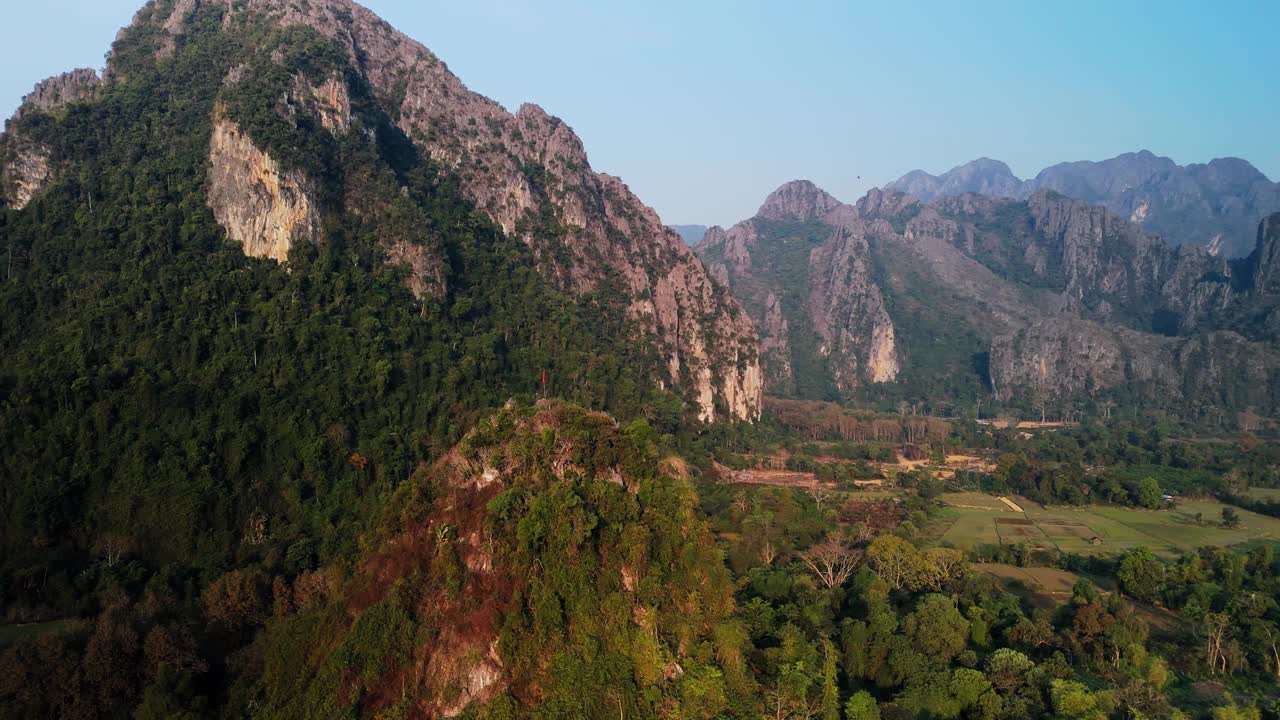 乡村的石灰岩岩层，山上有老挝国旗。Vang Vien山谷与冰川平原上的绿色农业用地。无人机空中旋转。