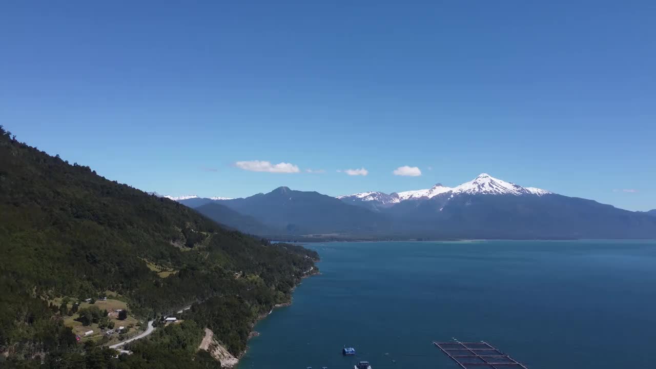 视频底部，巴塔哥尼亚的一个大湖上漂浮着一个大型养鱼场，背景相机从左到右平移着白雪皑皑的火山