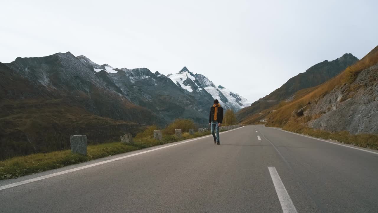 一名男子走在奥地利雄伟的大格洛克纳山路上，背景是积雪覆盖的尖峰和阿尔卑斯山脉的冰川。阿尔卑斯山的美景令人难以置信