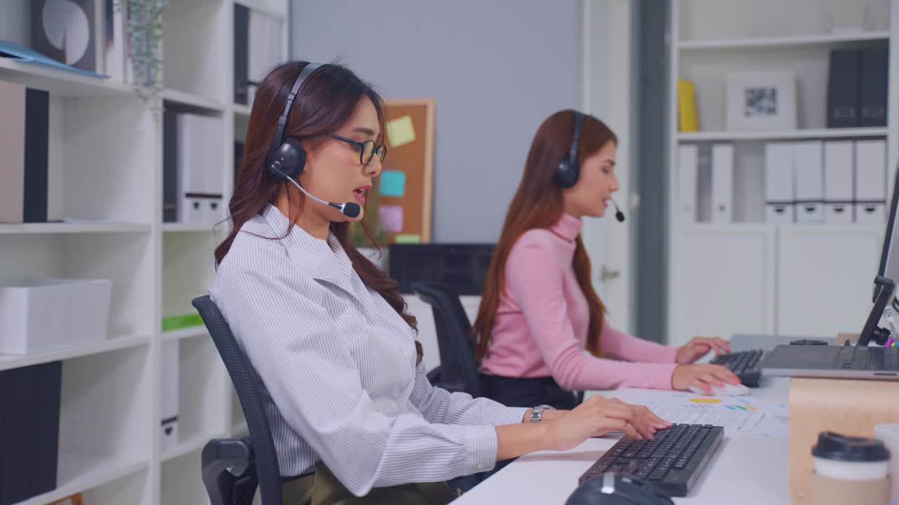 亚洲美丽的女商人在办公室公司的呼叫中心工作。有吸引力的年轻员工坐在桌子上，戴着耳机，使用笔记本电脑在工作场所支持客户。