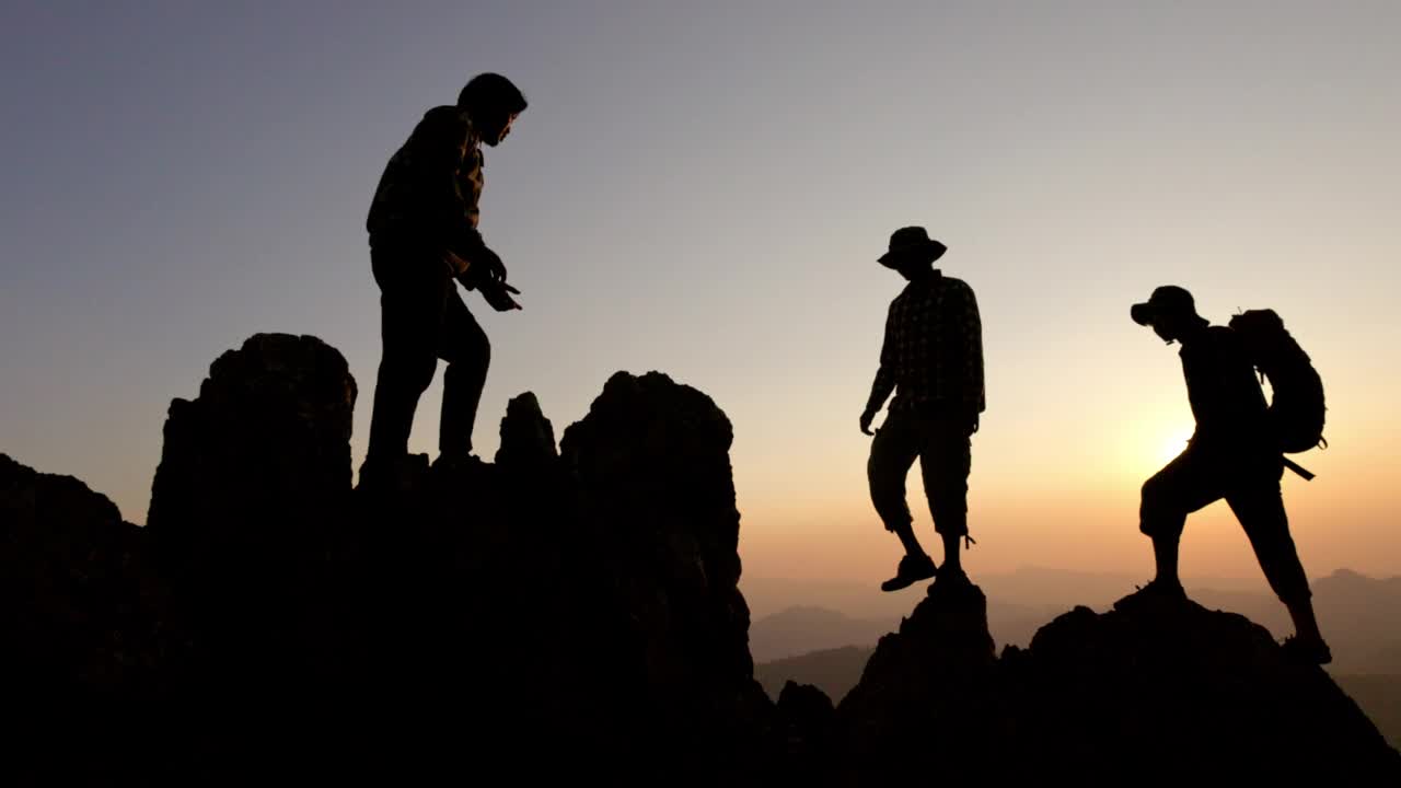 夕阳下徒步者互相帮助爬山的剪影。以人为本，注重团队合作。