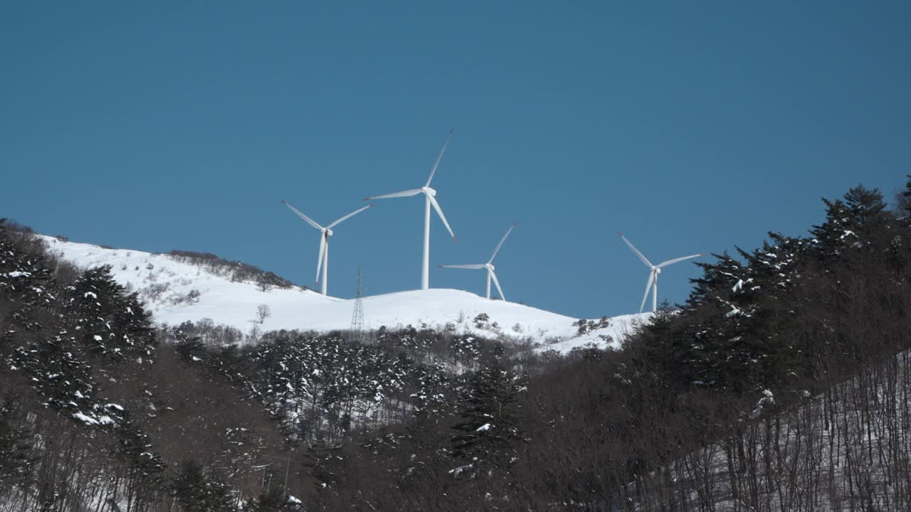 风力涡轮机旋转在白雪皑皑的山峰对蓝天与森林在前景低角度视图