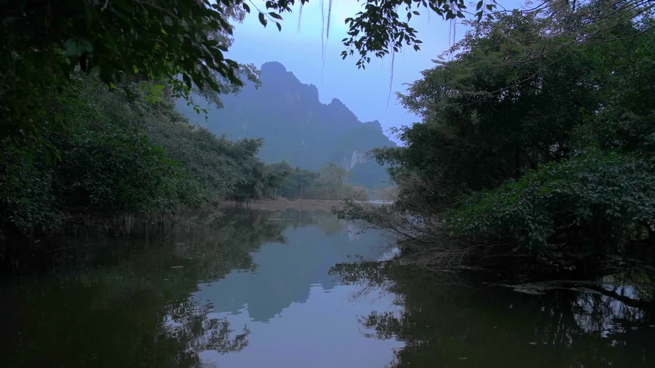 在阴天的越南宁平，小湖上茂密的树木植被反射出锯齿状山脉的剪影。远景
