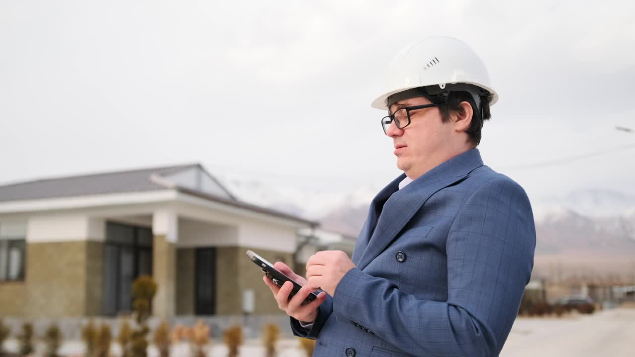 头戴白色安全帽的主管在建筑设备上用电话打字，近距离，侧面。