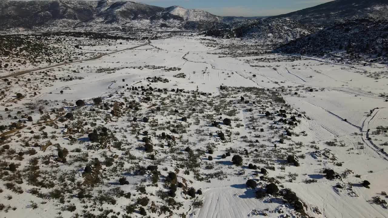 无人机的反向飞行聚焦于一个完全积雪的平原和灌木丛，我们想象了一条清除了积雪的土路，通往西班牙阿维拉的农村农场，背景是冬天的山脉