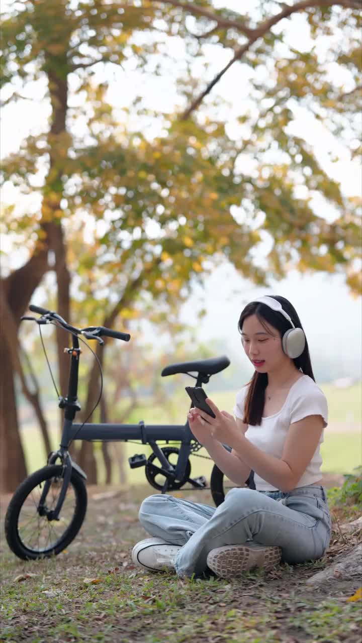 一名亚洲女子在公园骑自行车休息时，用手机选择并听她最喜欢的歌曲。在大自然中享受音乐