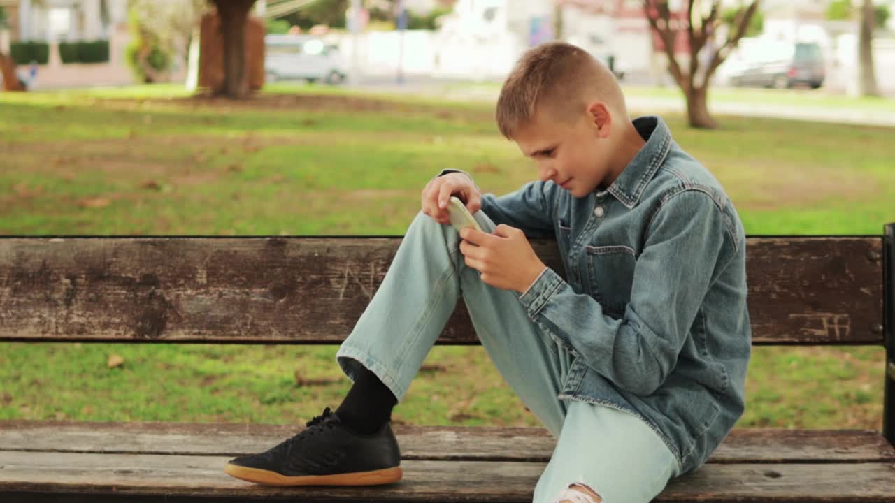 一个看起来像欧洲人的男孩坐在绿色城市公园的长椅上，一边在手机上输入信息，一边陷入沉思。说明现代通信的概念