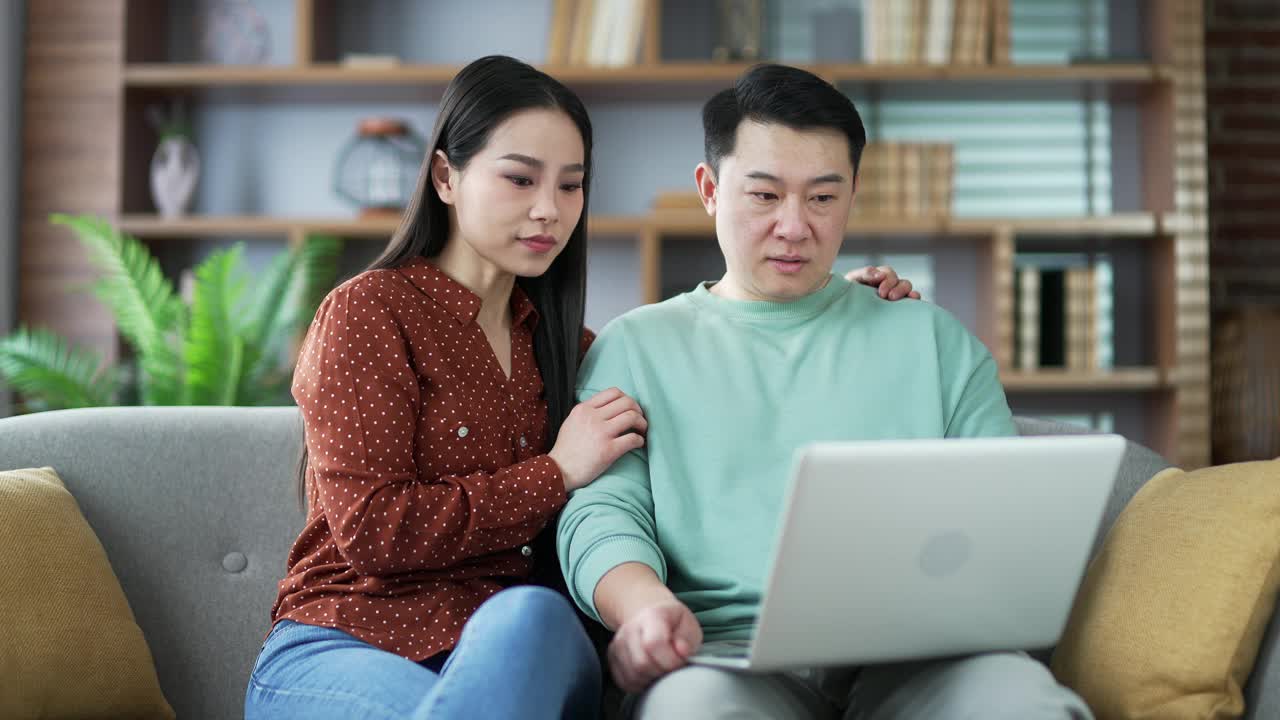 亚洲家庭夫妇坐在家里客厅的沙发上用笔记本电脑在网上购物时争吵。