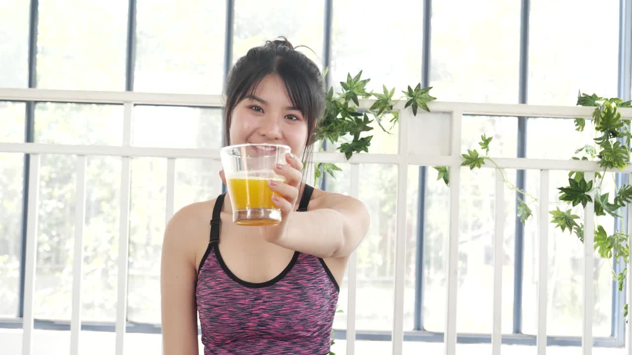 女人手里捧着冰凉的橙汁，新鲜的水果清凉地喝着。亚洲女性笑着笑着看镜头保健居家健身生活方式。美女从鸡尾酒瓶里倒橙汁