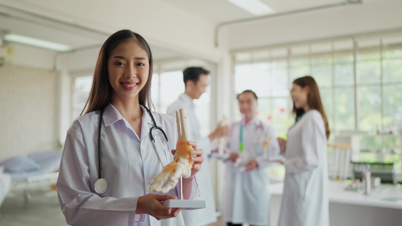 詳情圖-穿著制服的年輕亞洲女性骨科醫生的肖像，抱著人體踝關節骨骼模型，在醫院對著鏡頭微笑。醫療整形外科醫師