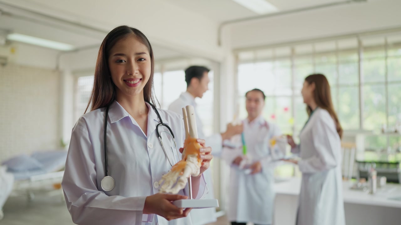 詳情圖-穿著制服的年輕亞洲女性骨科醫生的肖像，抱著人體踝關節骨骼模型，在醫院對著鏡頭微笑。醫療整形外科醫師