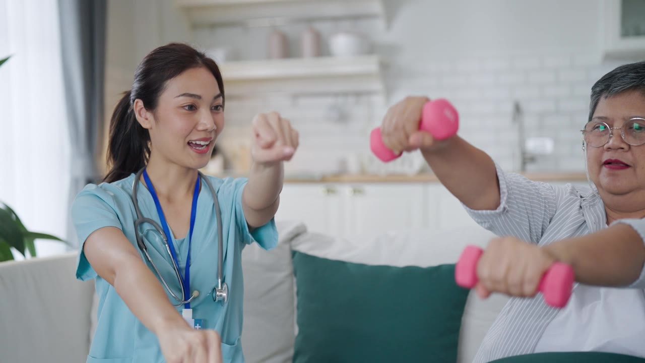 详情图-一位亚洲女性在家中使用哑铃进行手臂和手部理疗，理疗师在护士的帮助下进行康复治疗。