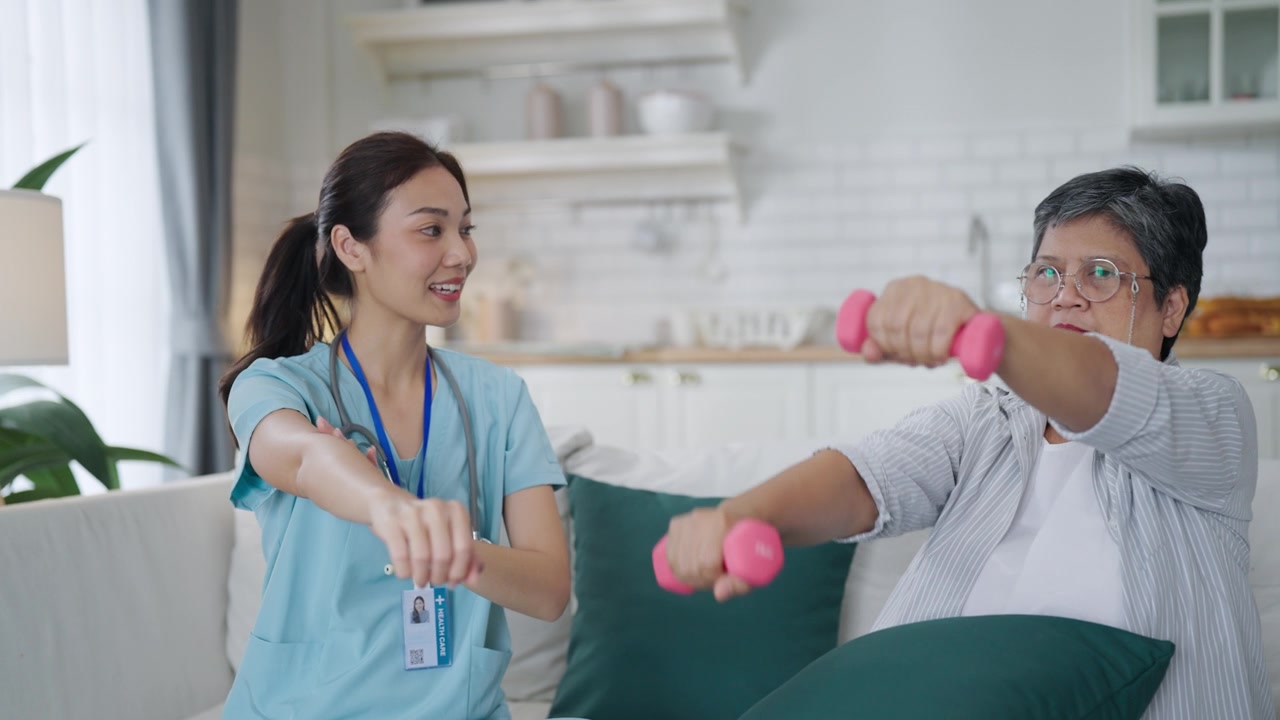 详情图-一位亚洲女性在家中使用哑铃进行手臂和手部理疗，理疗师在护士的帮助下进行康复治疗。