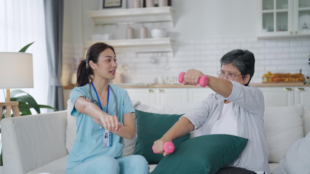 详情图-一位亚洲女性在家中使用哑铃进行手臂和手部理疗，理疗师在护士的帮助下进行康复治疗。