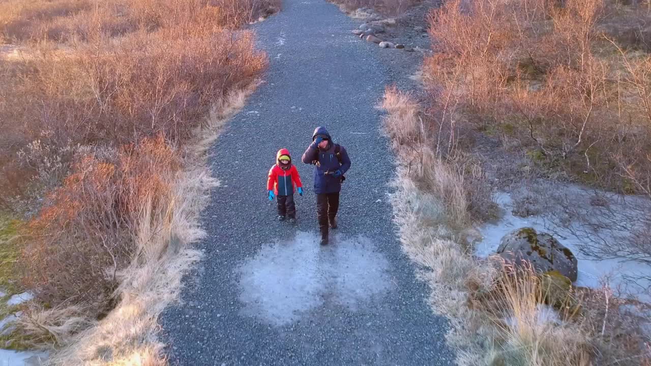 父子俩在冰岛Snaefellsjokull冰川附近徒步旅行的航拍照片，在金色的夕阳下，他们在贫瘠的土地上徒步旅行，在电影般的风景中向镜头挥手