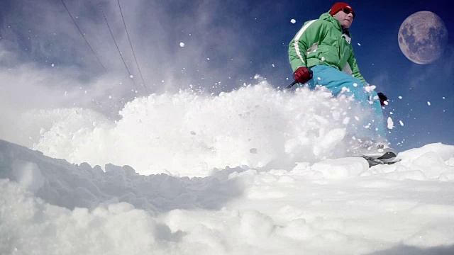 一名滑雪者在蓝天上有月亮的情况下从雪坡上滑下来的有力而充满活力的动态镜头。先是实时速度视频，然后减速来展示猛烈的雪暴。