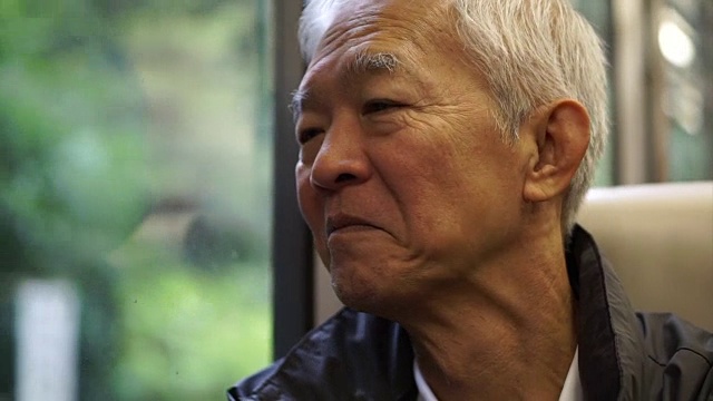 年长的亚洲人坐在火车上，透过窗户往外看。在退休旅行中思考和享受风景