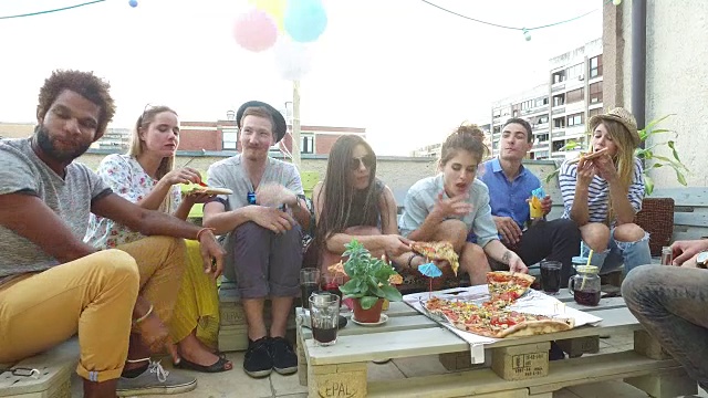 年轻人围坐在桌子旁，在屋顶露台上吃着披萨