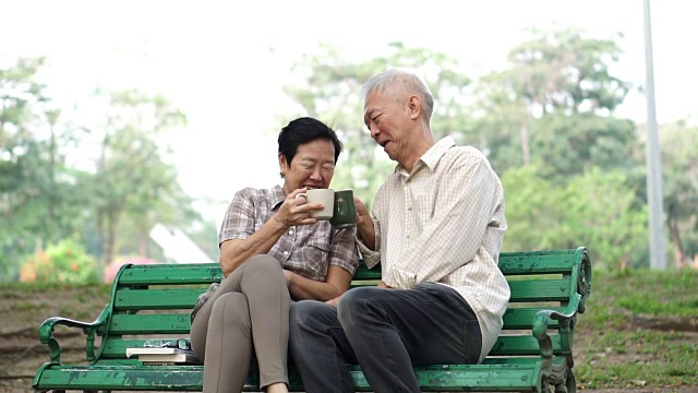 慢镜头快乐的亚洲老夫妇坐在早上的公园喝茶，喝咖啡。交谈并表达爱意