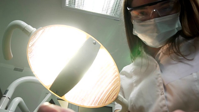 年轻女牙医戴着口罩，带着工具走近。牙科灯照进病人嘴里，站在病人身上，看着相机，牙医的脸