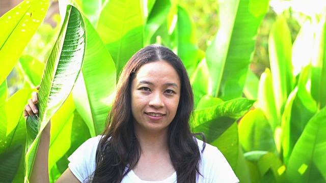 亚洲女人隐藏她的表情在热带树叶后面。微笑和快乐情绪管理理念