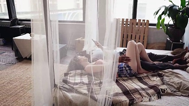 詳情圖-年輕的混血夫婦早上躺在床上聽著音樂。男人和女人使用智能手機(jī)，瀏覽互聯(lián)網(wǎng)