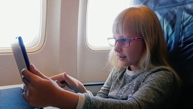 详情图-一个戴眼镜6年的女孩坐飞机旅行。在飞行中使用数字平板电脑