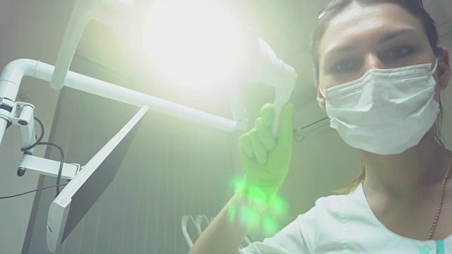 详情图-年轻的女牙医戴着面罩和防护手套，打开牙科灯，站在病人身上。看着摄像机，用4k拍的