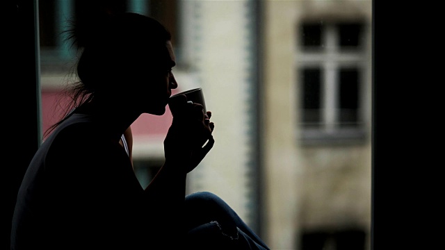 美丽的女人在窗前的特写肖像与一杯热饮在早上。坐在窗台上独自享受咖啡的神奇女孩剪影