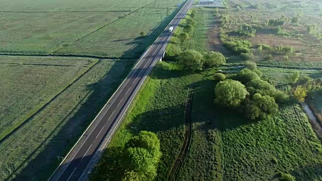 一辆卡车在太阳升起的绿色田野之间的道路上行驶的全高清航拍镜头