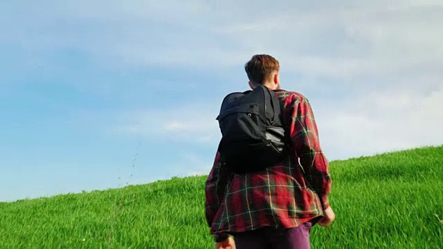 一个年轻人在爬一座绿色的大山。以蓝天为背景的风景景点的后视图。替身拍摄