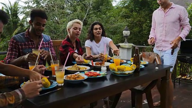 一名男子将食物传递给坐在餐桌旁的年轻朋友们，他们在夏季露台上聚会