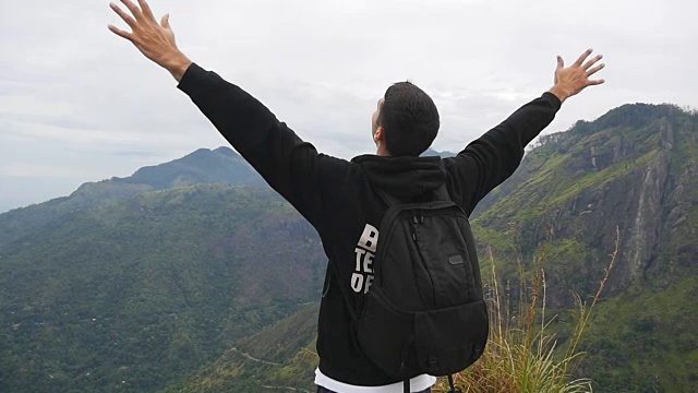 一名年轻男性徒步旅行者背着背包爬上山顶，举起双手。一名游客站在美丽的峡谷边缘，胜利地伸出双臂。慢镜头后视图