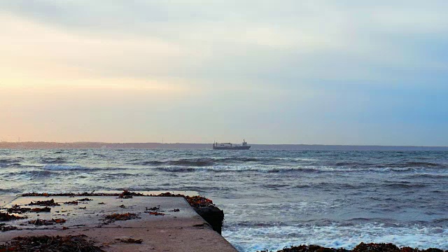 日落时海浪撞击码头的慢镜头，远处的货船在背景中