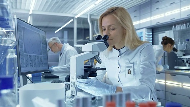 女性研究科学家在显微镜下观察生物样本。她和她的同事们在一家大型现代化实验室/医疗中心工作。