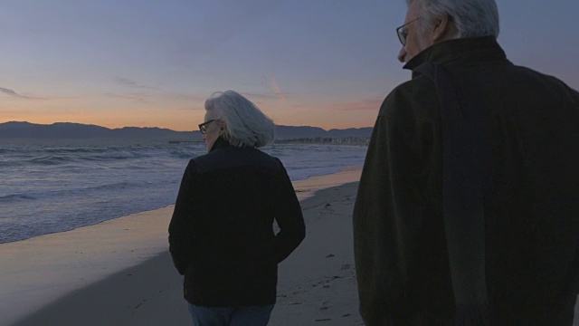 快乐的60多岁的老夫妇在夕阳下沿着高档海滨散步并指着