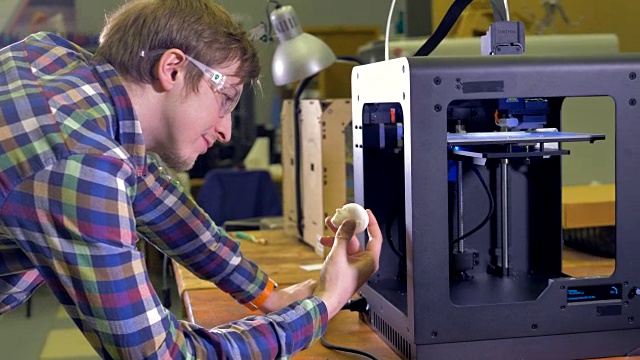 一个快乐的工程师拿着一个白色的头部原型，看着一台正在工作的3d打印机。4 k。