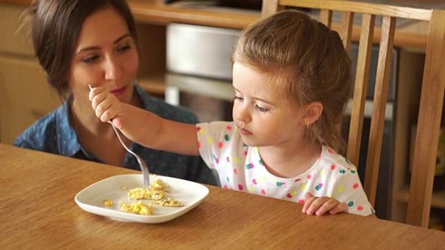 一个小女孩正在厨房里喂她的妈妈。他玩食物，把它掉在桌子上。