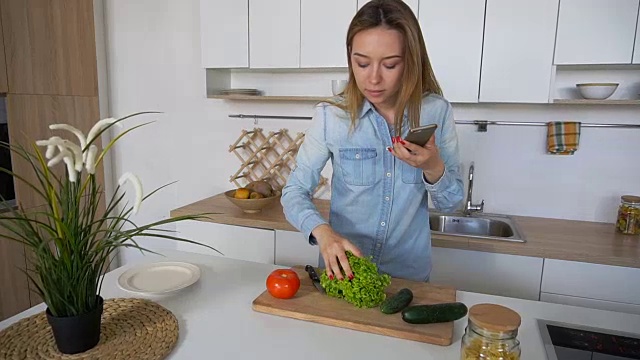 白天，迷人的女人站在时尚的厨房中央，手里拿着手机做饭，用蔬菜作图