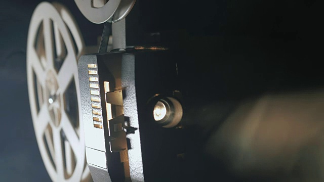 一个老式的古董超级8毫米电影放映机的正面视图，投射一束光在一个黑暗的房间旁边的一堆散开的电影