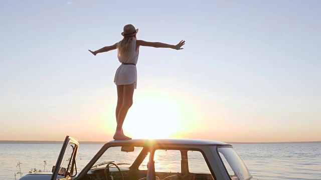 詳情圖-在慢鏡頭中，夕陽下的女子在背景海上的汽車上舉手，年輕女子站在車頂上，情感幸福，快樂的旅程
