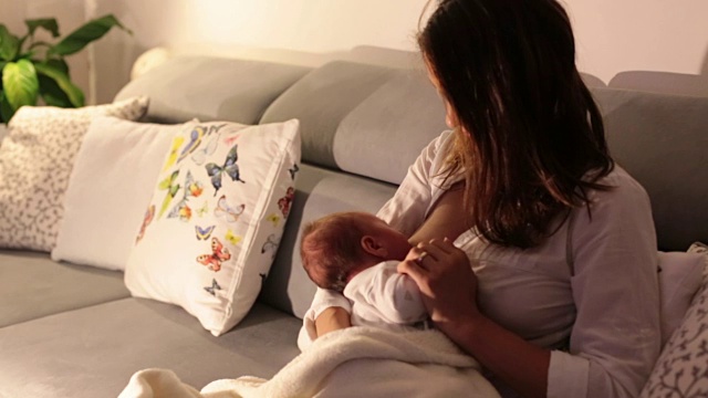 年轻美丽的母亲，在昏暗的灯光下给她的新生儿喂奶。妈妈母乳喂养婴儿